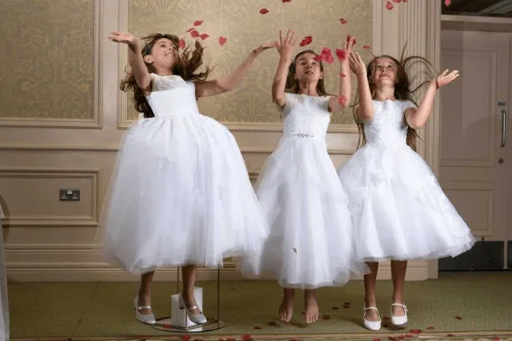 Three young girls in white dresses joyfully toss rose petals