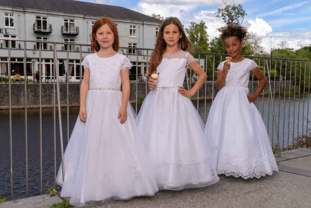 Three girls in elegant white dresses pose by a river, with one holding an ice cream cone.
