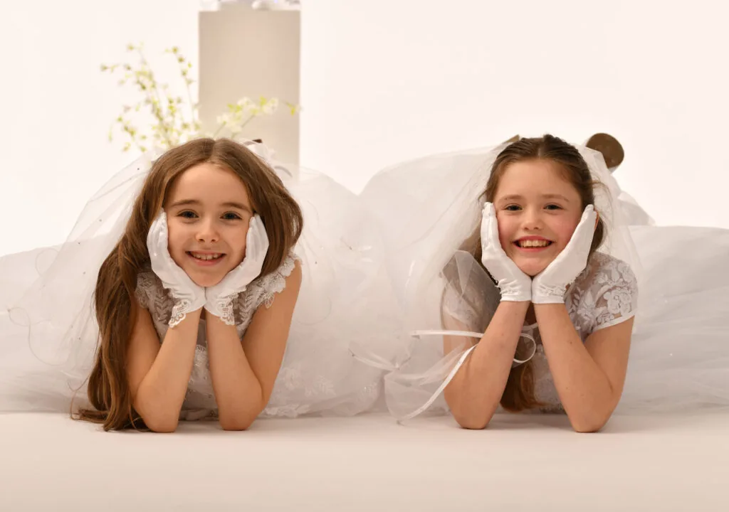 Two smiling young girls in white dresses, lying face down with hands on cheeks