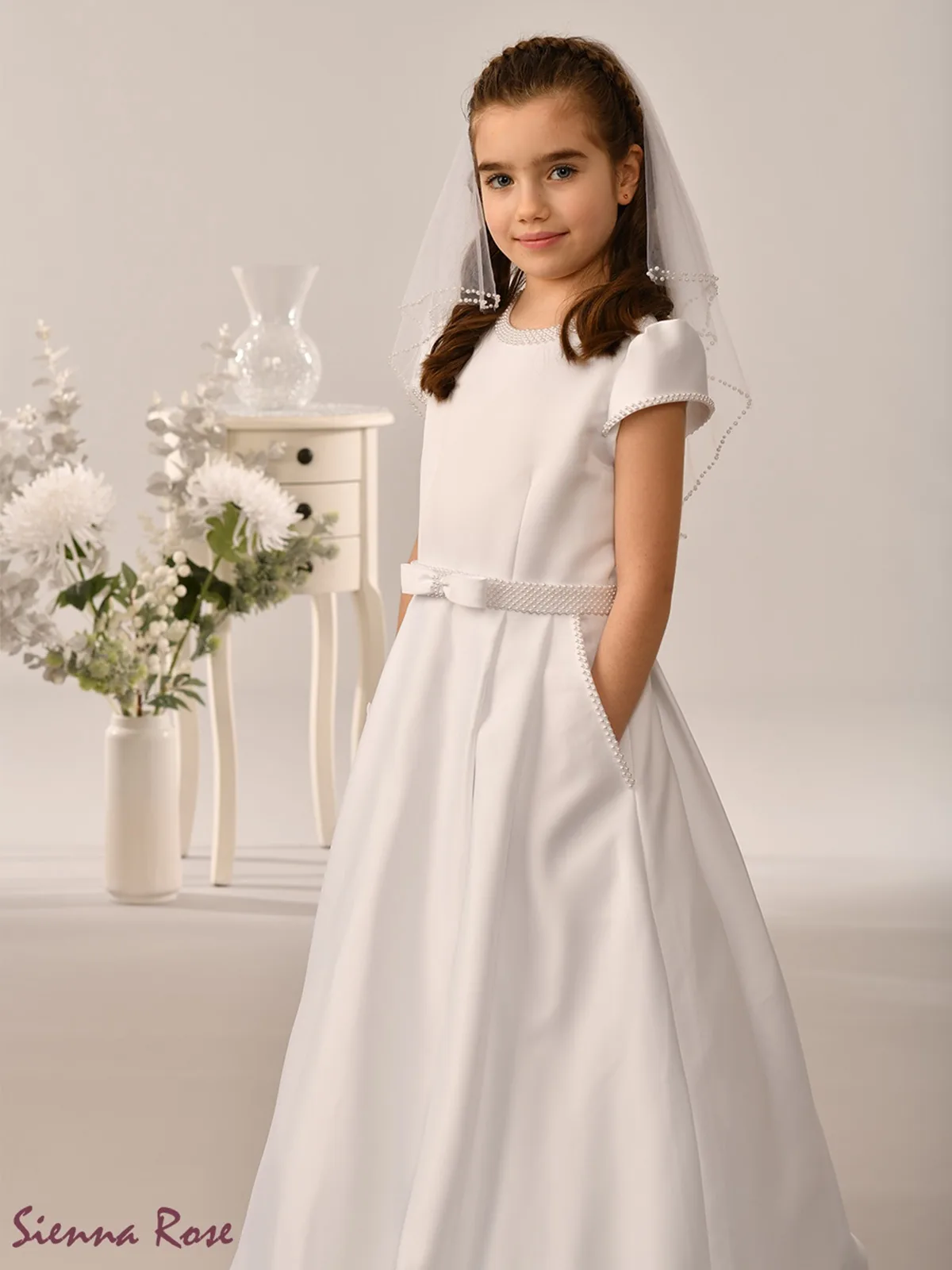 A young girl stands confidently in a white dress and veil, smiling gently.